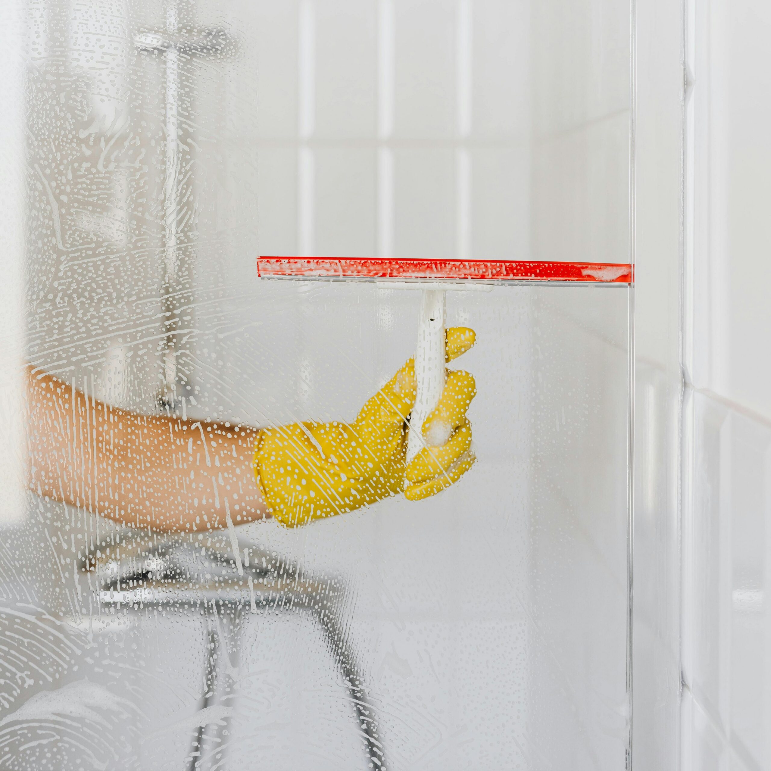 A person wearing a yellow glove cleans a shower glass with a squeegee, emphasizing hygiene and cleanliness.